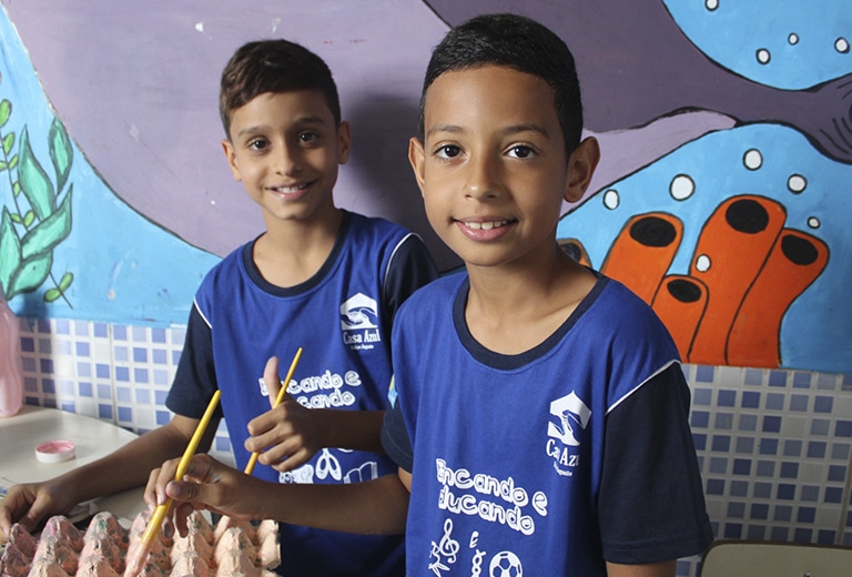 Casa Azul Dois meninos sorrindo com camisetas azuis da Casa Azul, segurando lápis durante atividade educativa, com mural colorido de fundo na instituição em Brasília.