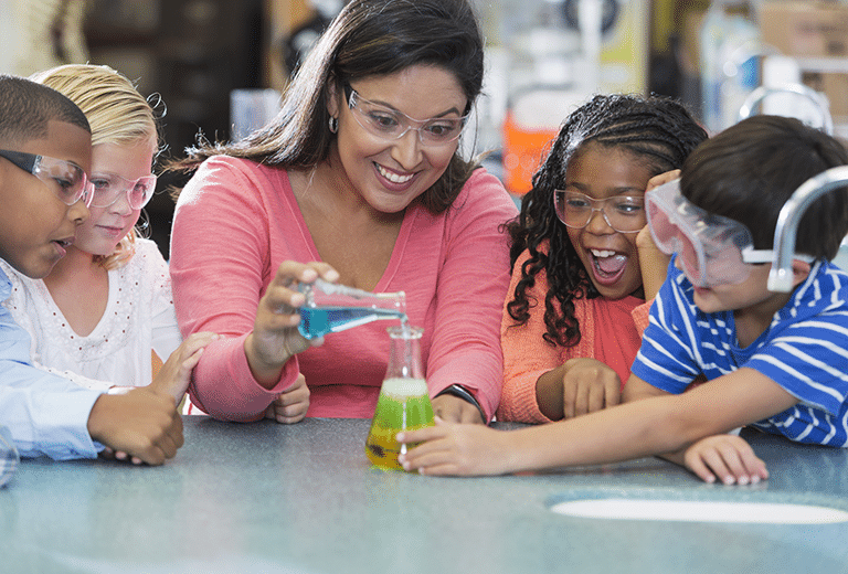 Dia Internacional da Mulher e da Menina na Ciência Uma professora orienta um pequeno grupo de crianças enquanto realizam um experimento científico simples. Usando óculos de proteção, elas observam com entusiasmo a mistura de líquidos coloridos em um frasco sobre a mesa, reagindo e mudando de cor.