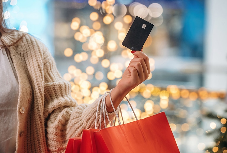 O melhor presente de fim de ano: equilíbrio financeiro Mão de mulher com unhas vermelhas segurando um cartão de crédito preto acima de uma sacola de compras laranja, com luzes douradas desfocadas e árvore de Natal ao fundo.